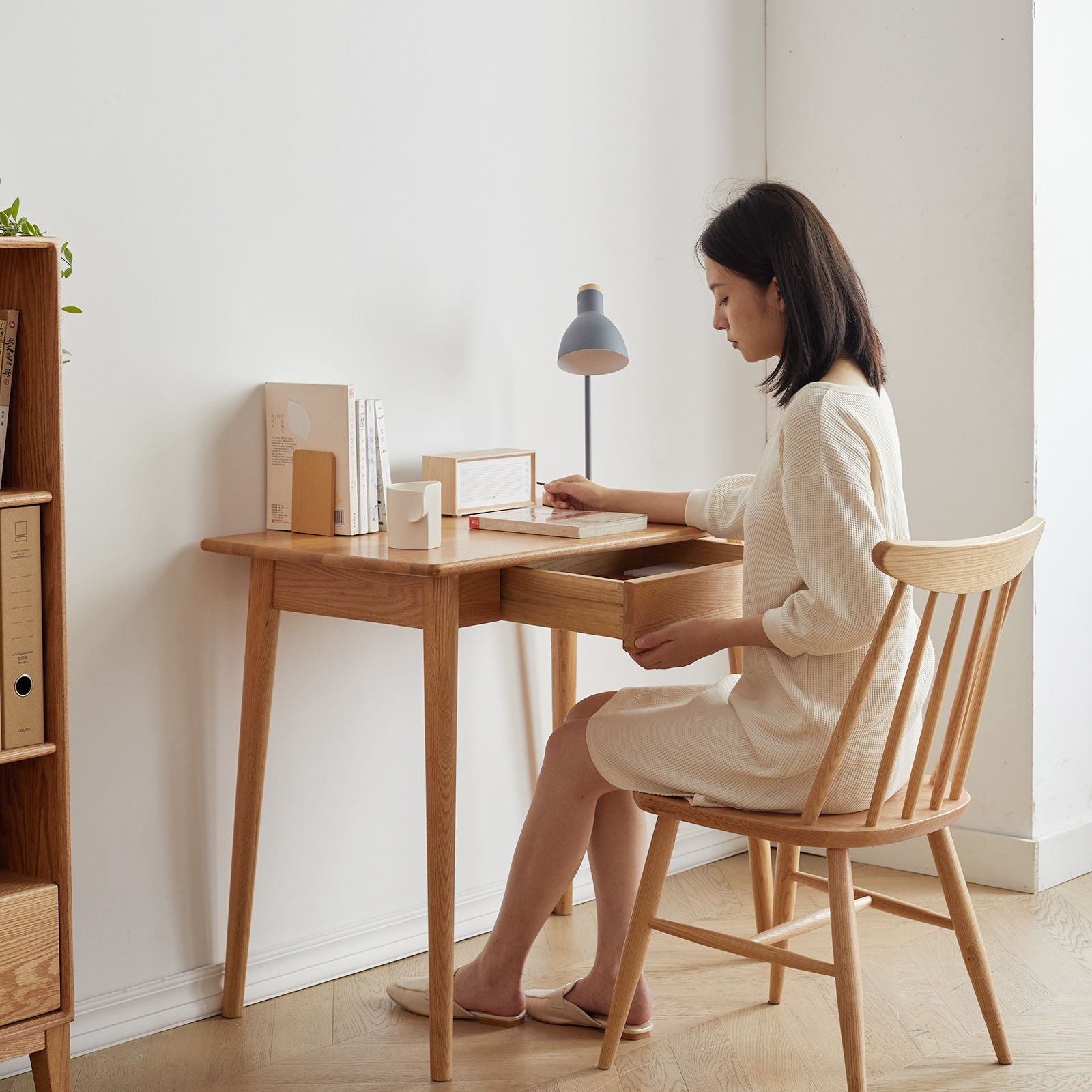 A woman in a cream dress sits at an oak natural computer desk with a drawer, facing sideways near a bookshelf. A small lamp and books rest on the desk, creating a softly lit, minimalist, and serene atmosphere.