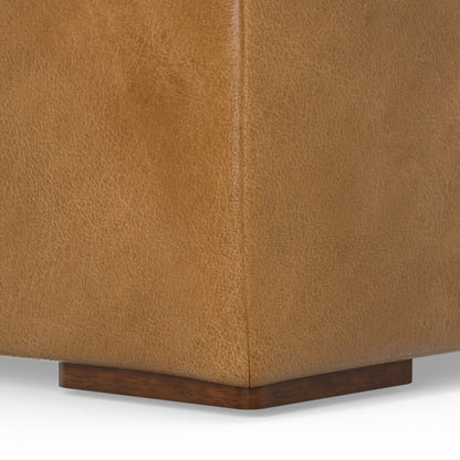 Close-up of a light brown leather sofa corner showing the textured surface and wooden base. The material has a slightly distressed look, and the wooden base appears polished and dark in contrast with the leather.