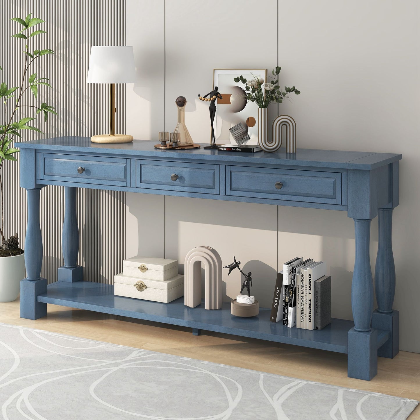 The Long Console Table with a blue design, three drawers, and a lower shelf is set against a wall. It displays decor, books, and a potted plant. A modern lamp sits on top, while below more items are displayed. The floor features a light rug with circular patterns.