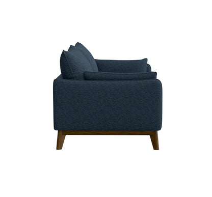 Side view of the Varia - Sofa - Blue, a modern dark blue upholstered piece with wooden legs, featuring a boxy design and plush cushions for seating and back support, set against a plain white background.