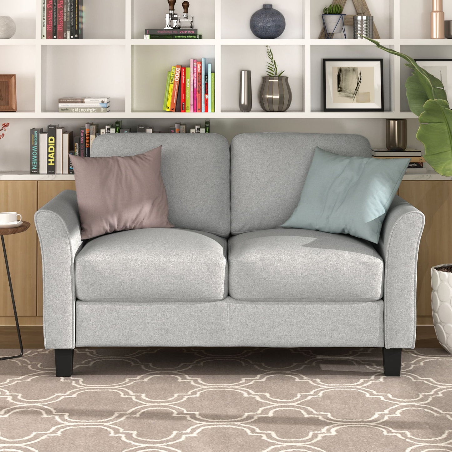 A cozy living room features a light gray love seat sofa with pink and blue cushions. Behind it sits a bookshelf filled with books, decorations, and plants. A large green leaf plant is visible to the right, complemented by a geometric patterned rug.