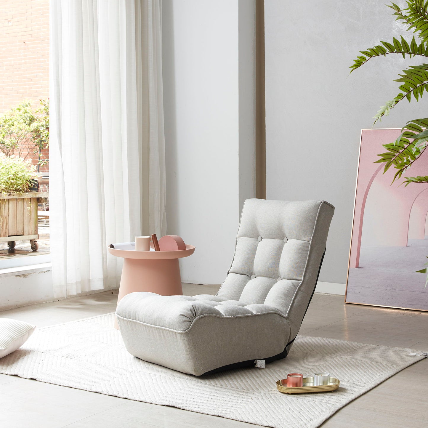 A cozy, light-gray Reclining Japanese Chair Lazy Sofa sits on a textured rug near a window. Beside it is a small pink table with trays holding items. A leafy plant and framed art with pastel colors lean against the wall, enhancing the serene atmosphere.