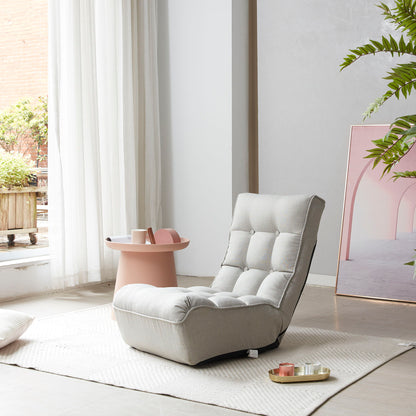 A cozy, light-gray Reclining Japanese Chair Lazy Sofa sits on a textured rug near a window. Beside it is a small pink table with trays holding items. A leafy plant and framed art with pastel colors lean against the wall, enhancing the serene atmosphere.