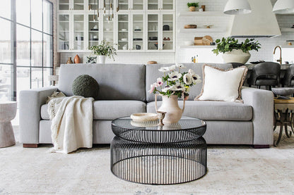 Modern living room featuring the Barton Sofa in gray, embellished with cushions and a throw blanket. In front, a glass coffee table is accessorized with vases of flowers. The backdrop includes open shelves and a kitchen area illuminated by natural light from a large window.