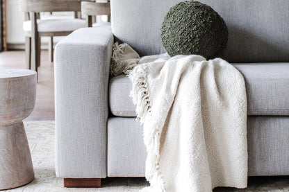 A Barton Sofa in light gray is adorned with a textured dark green cushion, and a soft, cream-colored throw is elegantly draped over one arm. Nearby, a small round wooden side table complements the setup, while a dining area with chairs can be seen in the background.