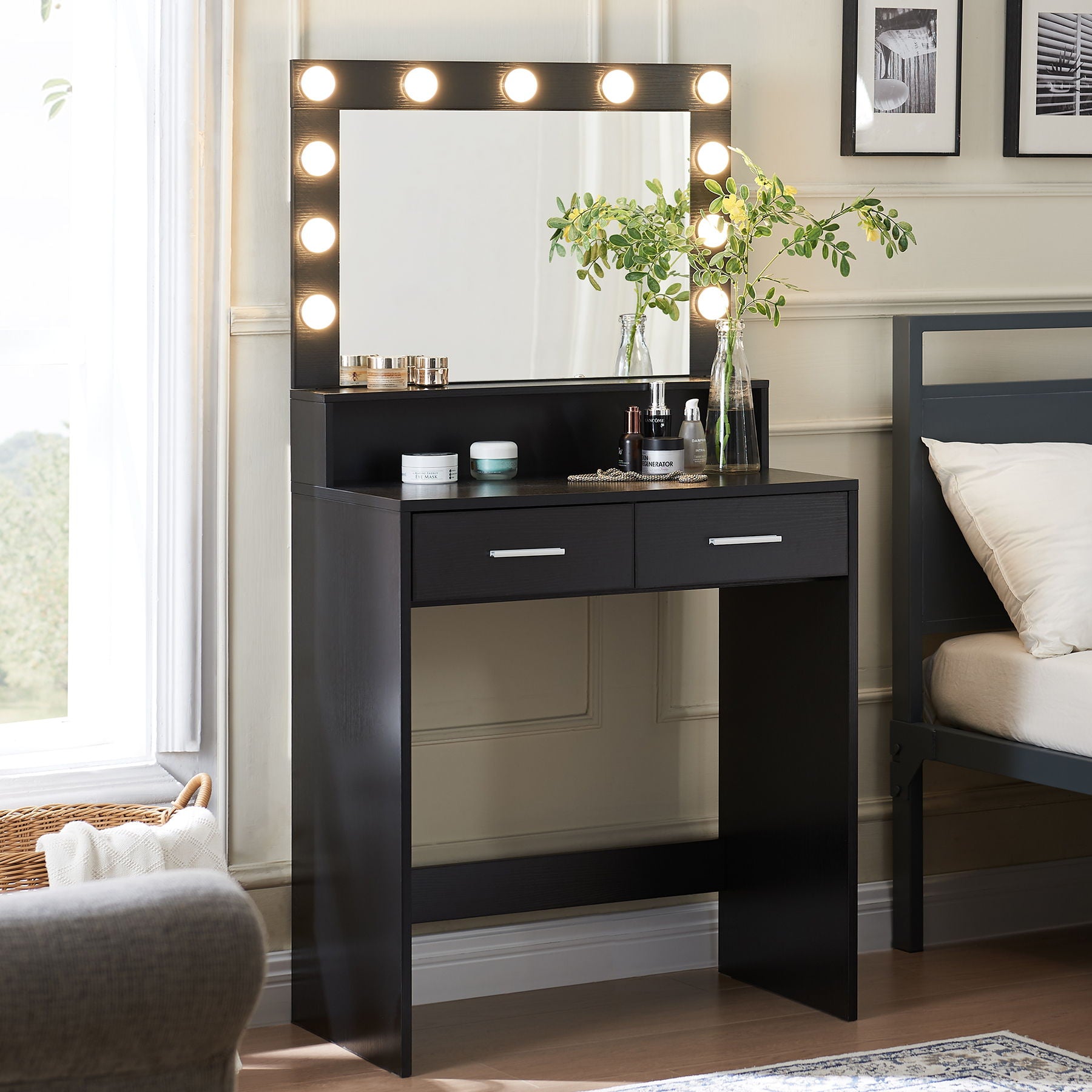 A black Vanity Desk With Mirror And Lights, featuring a large drawer and adjustable lighting, stands in a bedroom. Cosmetics and greenery decorate the table. Light-colored walls surround the space, with parts of a bed and sofa visible.