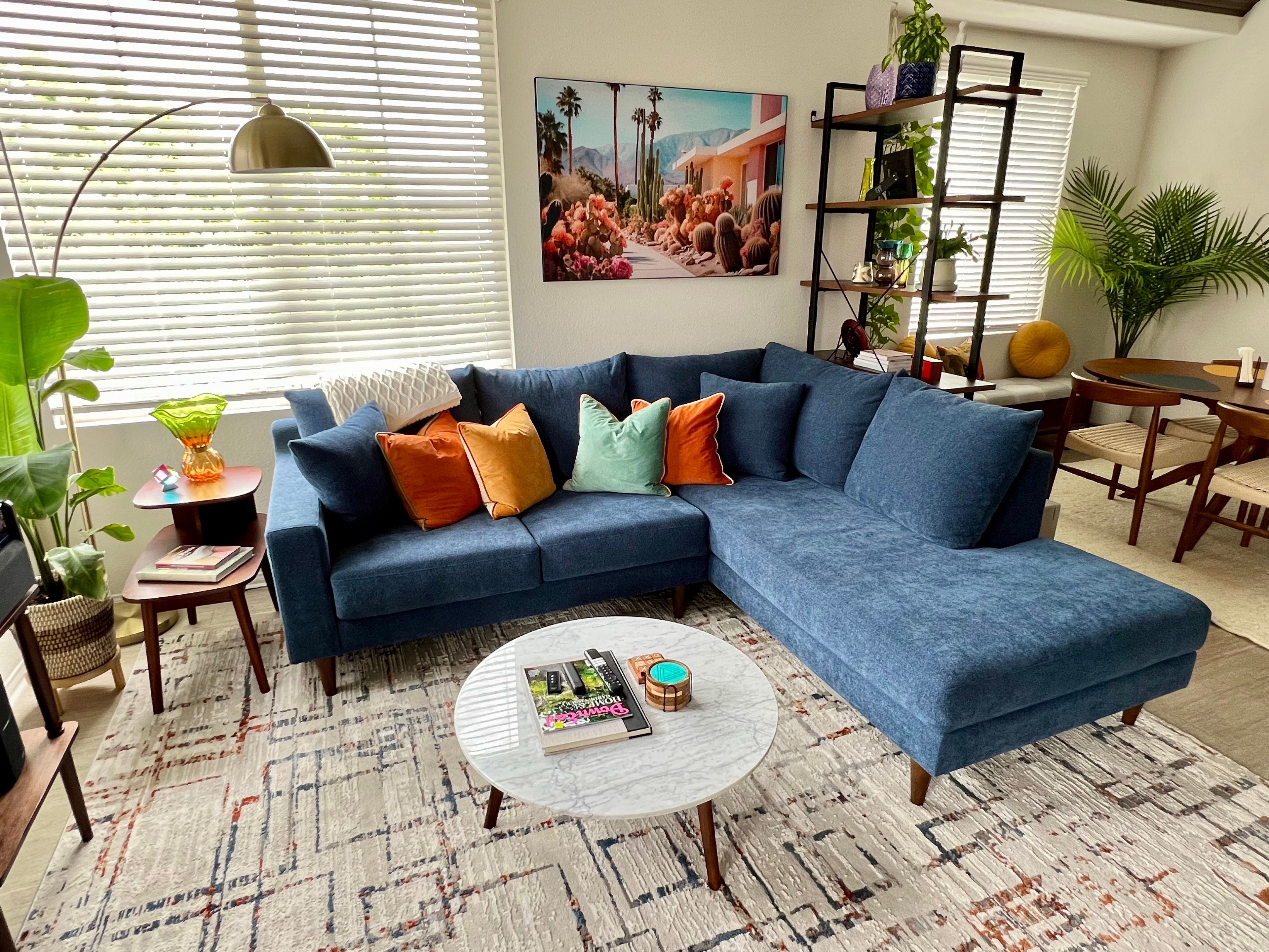 A modern living room with a blue sectional sofa adorned with colorful pillows. A round marble coffee table holds magazines and a small candle. Green plants, a floor lamp, and a scenic wall art piece add to the decor. Light streams in through white blinds.