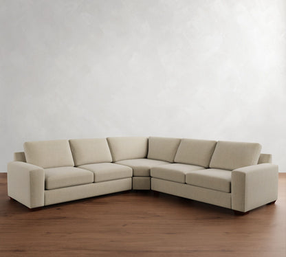 A Big Sur Square Arm Upholstered 3-Piece L-Shaped Wedge Sectional in beige is arranged on a wooden floor against a light gray wall. The sectional features three cushioned sections with seats and backrests, forming a cozy corner seating arrangement.