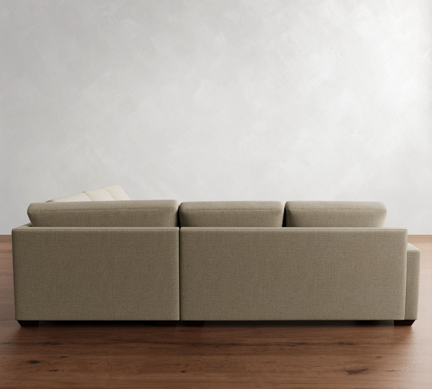 The Big Sur Square Arm Upholstered 3-Piece L-Shaped Wedge Sectional, with its minimalist beige design, low backrest, and blocky contours, is placed on a wooden floor. Its simple and modern style stands out against the textured white wall in the background.