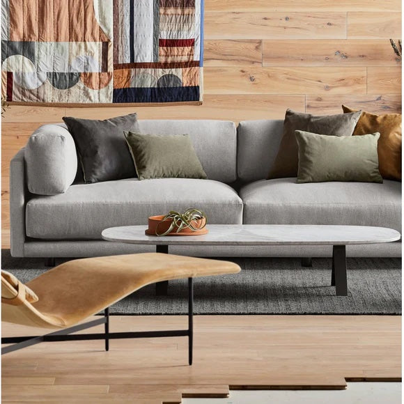 A modern living room featuring a light gray sofa with green and brown throw pillows, a low white rectangular coffee table with a plant on it, and a beige lounge chair. The wooden wall has decorative fabric art, and the floor is made of light wood.