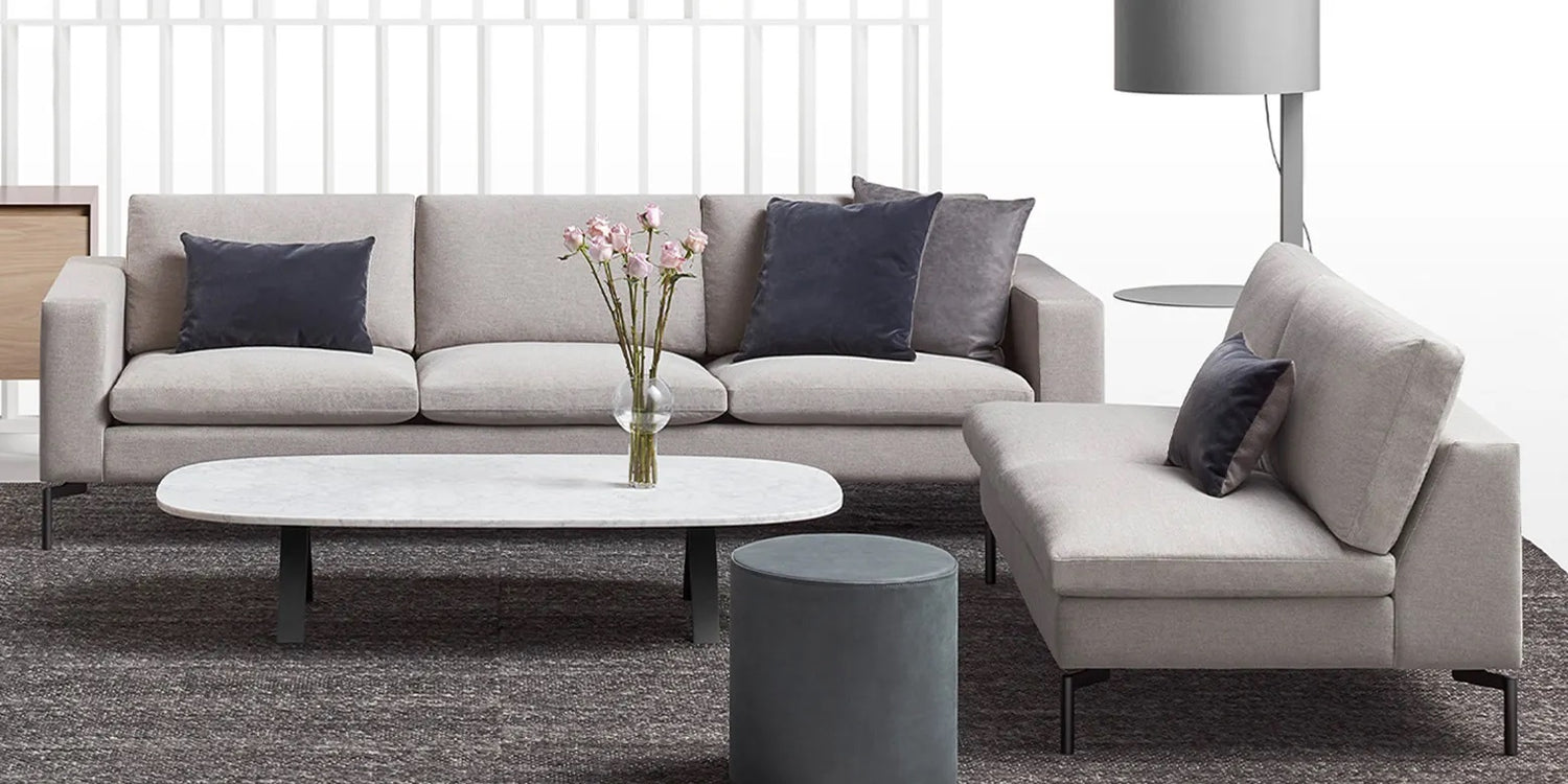 A modern living room features a large gray sectional sofa with several dark gray and light gray cushions, a white marble coffee table with pink flowers in a vase, a round gray ottoman, and a floor lamp. The furniture sits on a dark textured rug.