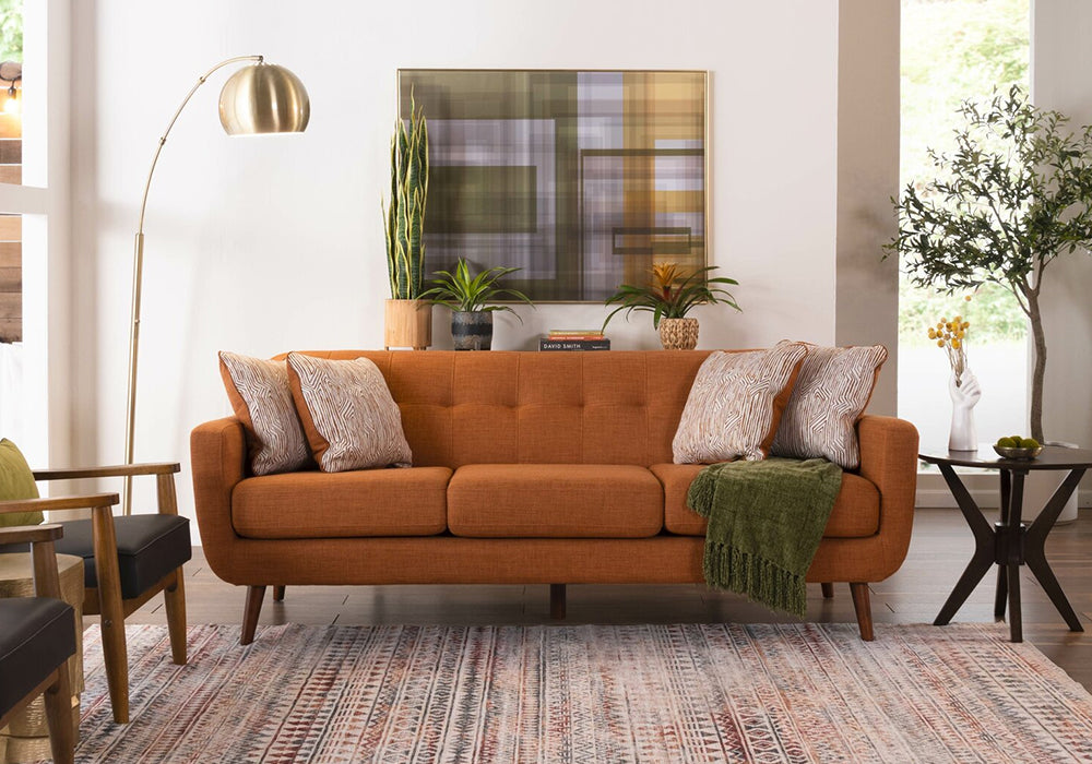 A cozy living room features an orange mid-century modern sofa adorned with patterned throw pillows and a green blanket. A floor lamp arches over the sofa, and a potted plant rests beside it. Abstract artwork and various plants decorate the space.