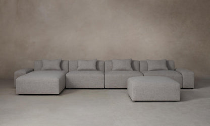 The Breuer Modular Chaise Armless Sectional 114" in Belgian Linen Agate, a spacious and gray-toned modular sectional sofa with multiple cushions, is arranged on a neutral-toned floor against a plain beige wall. The setup includes a matching ottoman positioned slightly to the right.