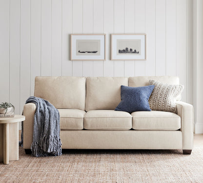 A Buchanan Square Arm Sleeper Sofa in beige is adorned with a blue pillow and a chunky knit pillow, positioned against a white paneled wall. Draped over one arm is a blue throw blanket. Above the sofa are two minimalist framed artworks, while to the left stands a small round table with a plant.