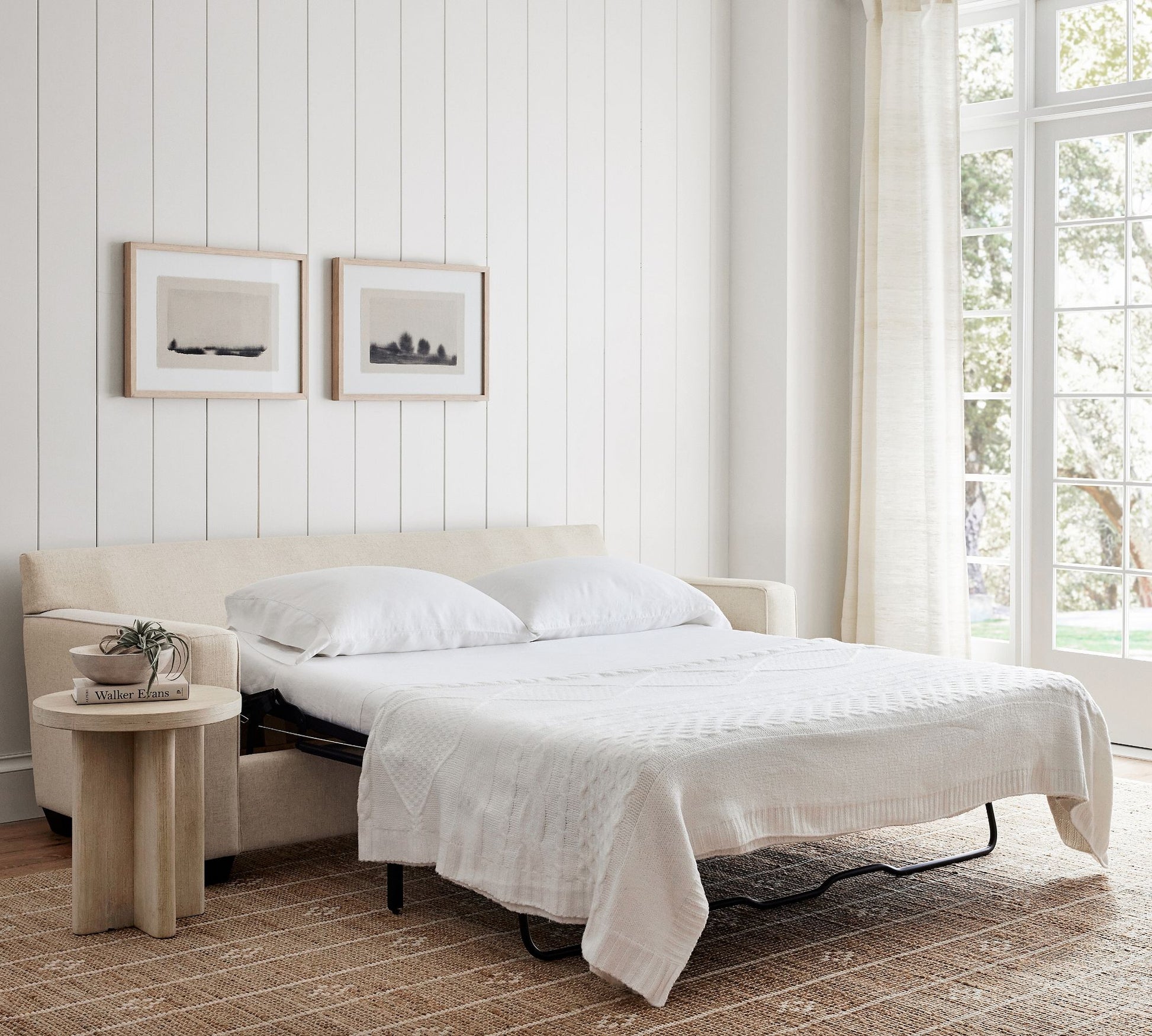 A cozy bedroom features the Buchanan Square Arm Sleeper Sofa, transformed into a bed and draped with crisp white bedding. Above it, two minimalist paintings adorn the wall. Nearby, a small round wooden side table is stacked with books and supports a glass. Large windows provide an expansive view of the greenery outside.