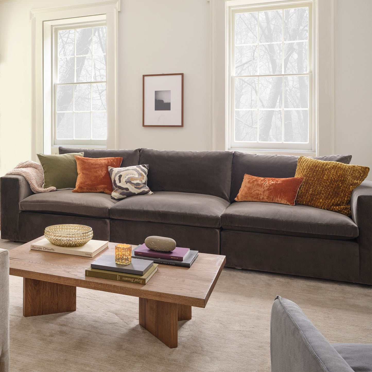 A cozy living room is centered around the Harmony Modular 3 Piece Sofa in dark gray, embellished with colorful throw pillows. In front stands a wooden coffee table showcasing books and various decorative items. Light streams through two large windows, while a framed picture adds brightness to the space, complemented by a soft carpet that completes the setting.