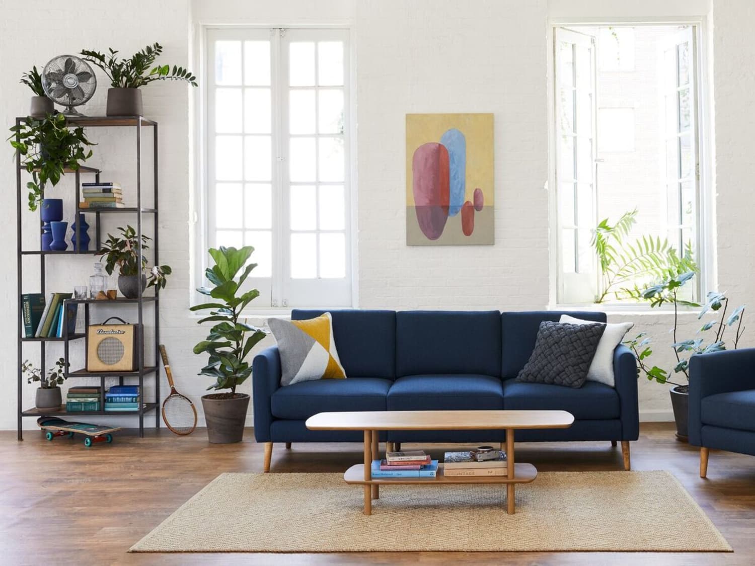 A modern living room with a navy blue couch, a wooden coffee table, and various potted plants. There's a geometric painting on the white brick wall between two large windows. A shelving unit holds books, decor, and a metal fan. The floor is wooden.