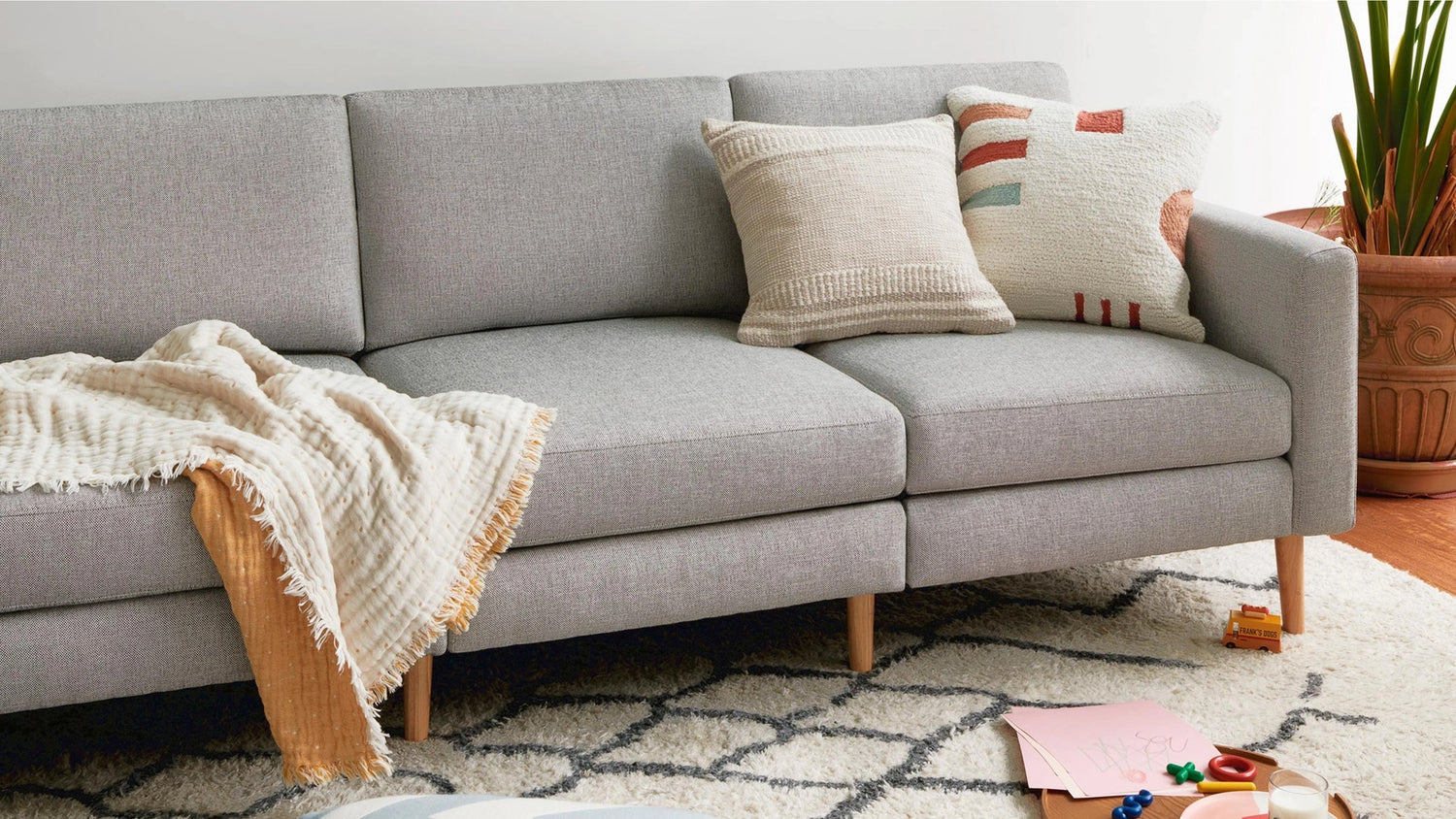 A modern gray fabric sofa is adorned with two decorative pillows and a cozy throw blanket. The couch sits on a patterned rug with geometric designs. A large potted plant is in the corner, and some children's toys and papers are scattered on the floor in the foreground.