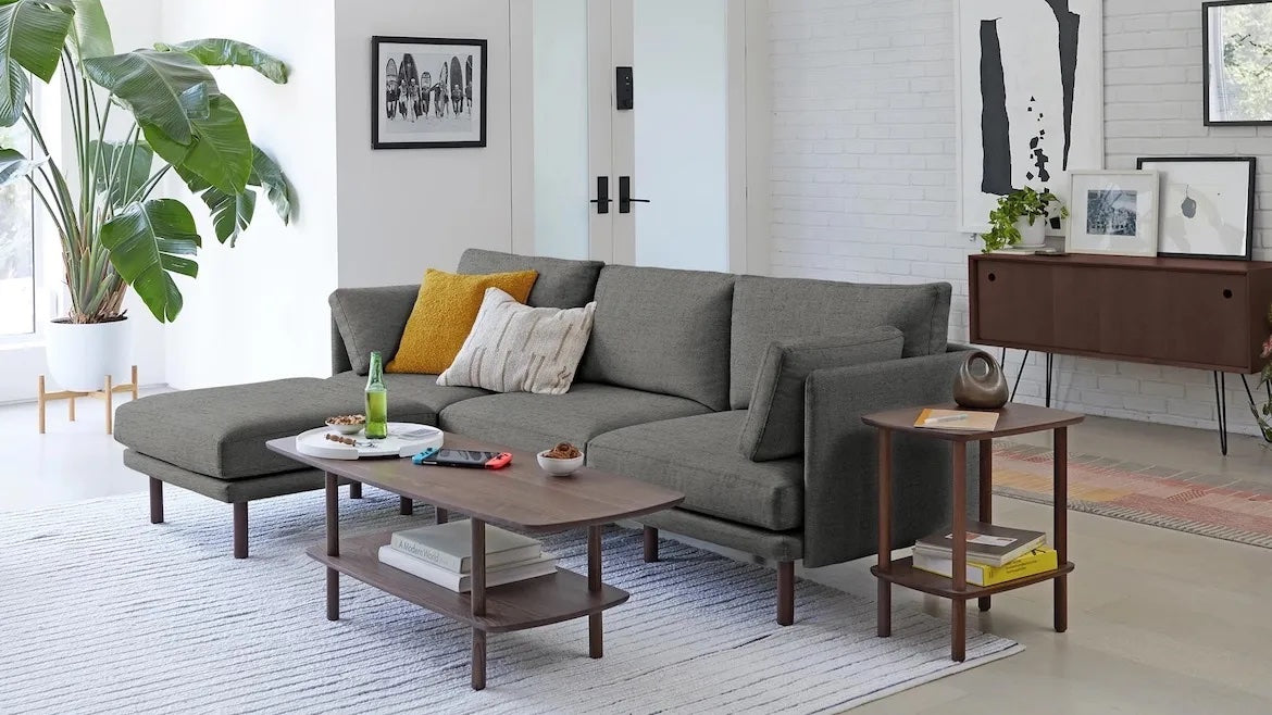 A modern living room features a gray sectional sofa with multiple cushions, a wooden coffee table and side table, a potted plant, and artwork on white brick walls. Natural light from large windows illuminates the space, highlighting the minimalist decor.