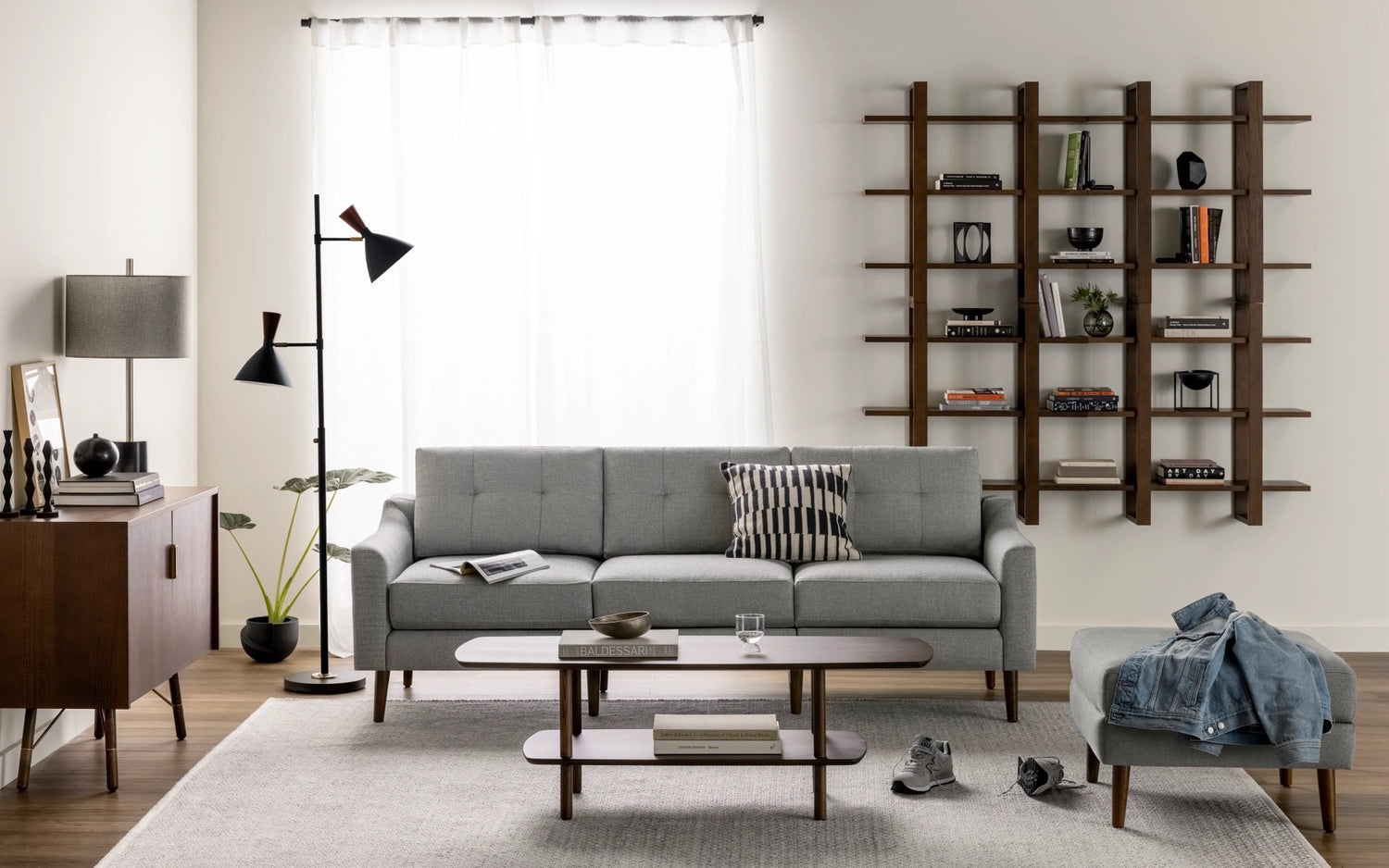 A modern living room featuring a light gray sofa with a striped pillow, a wooden coffee table, a blue armchair with a denim jacket, a wooden cabinet with a lamp, and a floor lamp. A wall-mounted bookshelf holds books and decor. Large window with sheer curtains.