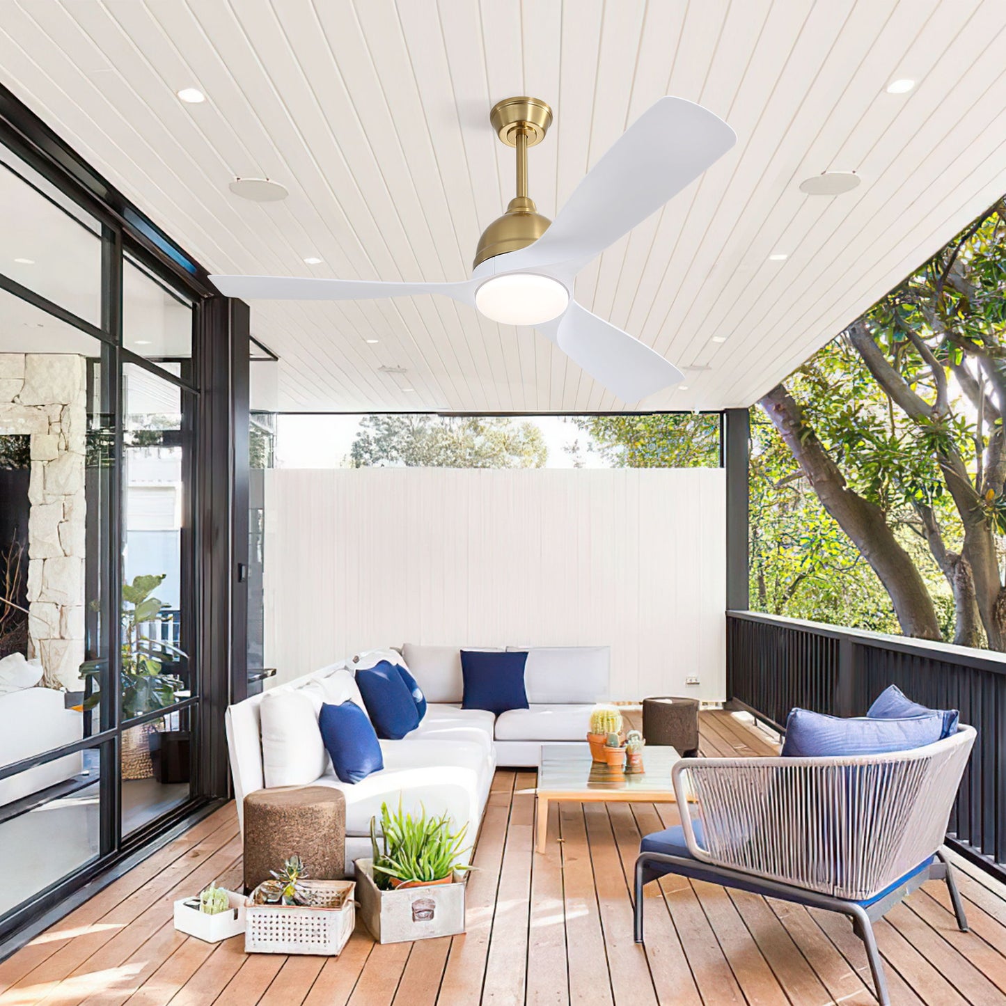 A modern outdoor living area with wooden flooring and a white ceiling showcases a white sectional sofa with blue cushions, a wicker chair, plants, and a coffee table. It includes large windows revealing trees outside and features the Modern Ceiling Fan with Smart App Control.