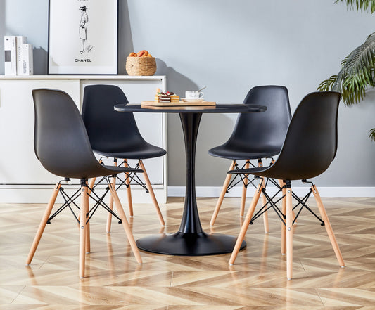 A modern dining area showcases a black MDF pedestal table from the 5-piece set for 4-6 people, accompanied by four matching chairs with wooden legs. The space features light wood flooring, a coffee cup and books on the table, a corner plant, and framed artwork on white surfaces.