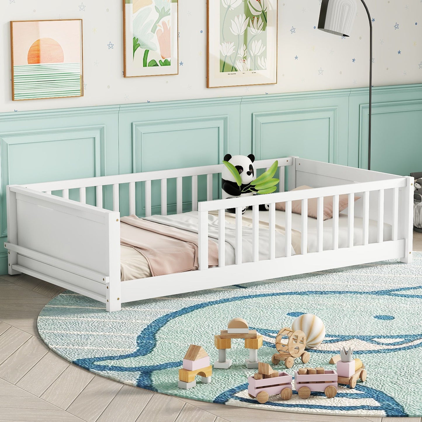 A floor platform bed with built-in book storage rack and white rails is in a room with mint green paneling, featuring a panda toy and pastel bedding. A large round bear-design rug on the floor is surrounded by wooden toys, while floral and sunset art adorns the walls.