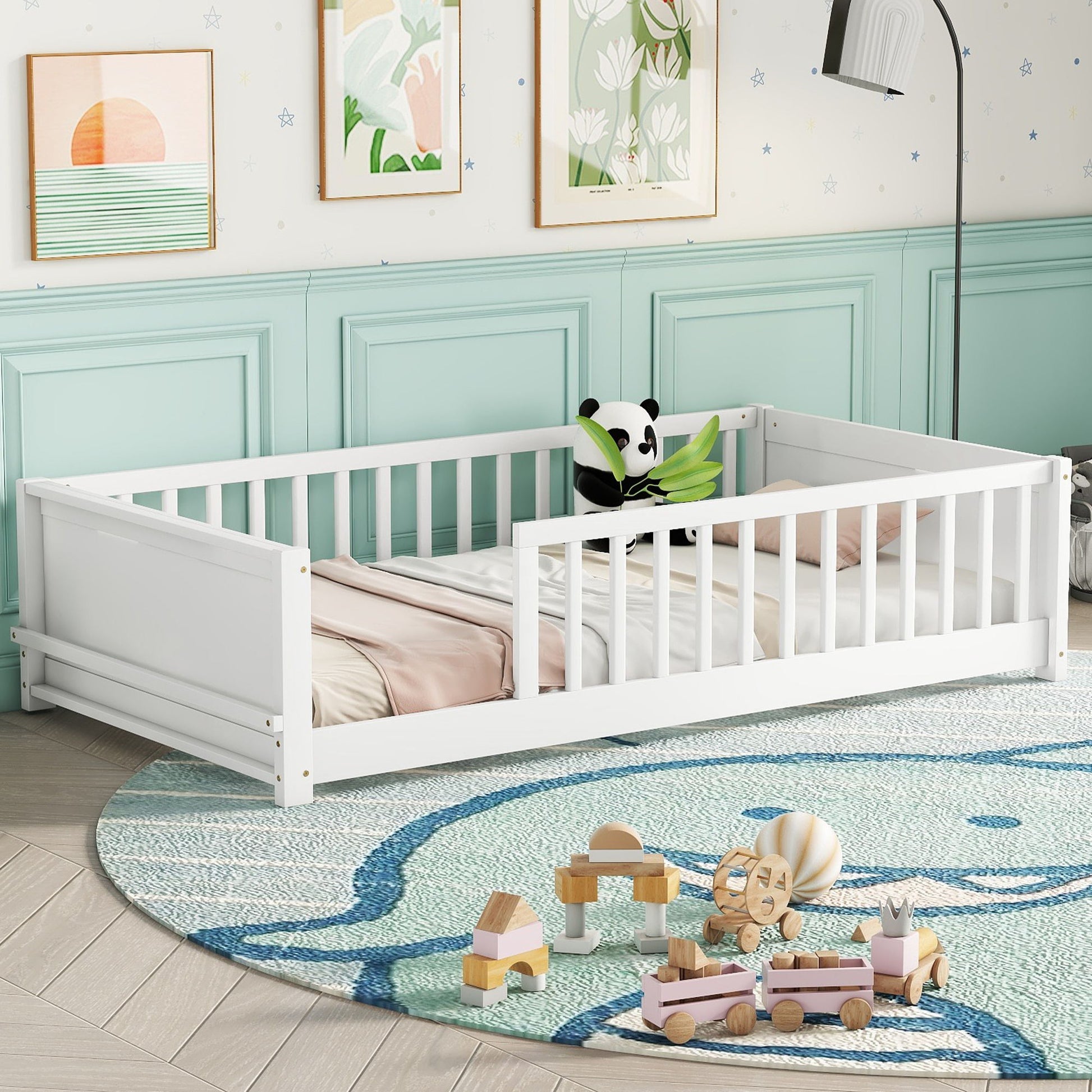 A floor platform bed with built-in book storage rack and white rails is in a room with mint green paneling, featuring a panda toy and pastel bedding. A large round bear-design rug on the floor is surrounded by wooden toys, while floral and sunset art adorns the walls.