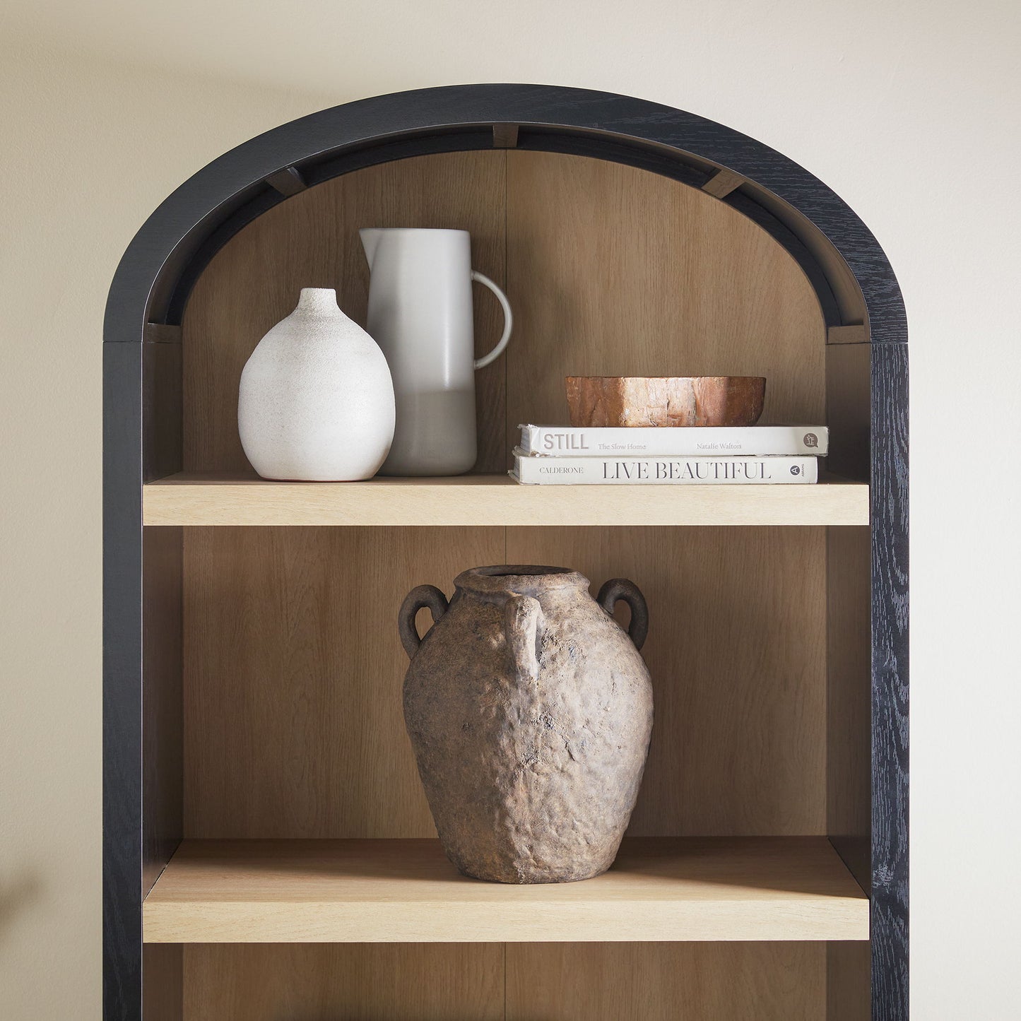 The Modern 5 Shelf Open Arched Bookshelf features a black frame and elegantly displays a white vase, gray pitcher, books, and a small wooden bowl on the top shelf. A large rustic clay jug adorns the middle shelf.