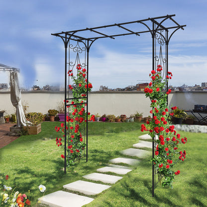 A garden showcases the Metal Garden Arch in black, supporting vibrant red roses climbing its sides. A stone path winds through lush grass to the arch, surrounded by potted plants and beneath a clear blue sky.