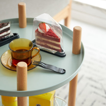 On a small round table with wooden legs, two black trays hold chocolate and red-layered cake slices. A transparent cup with amber liquid sits on a yellow saucer beside a black fork.