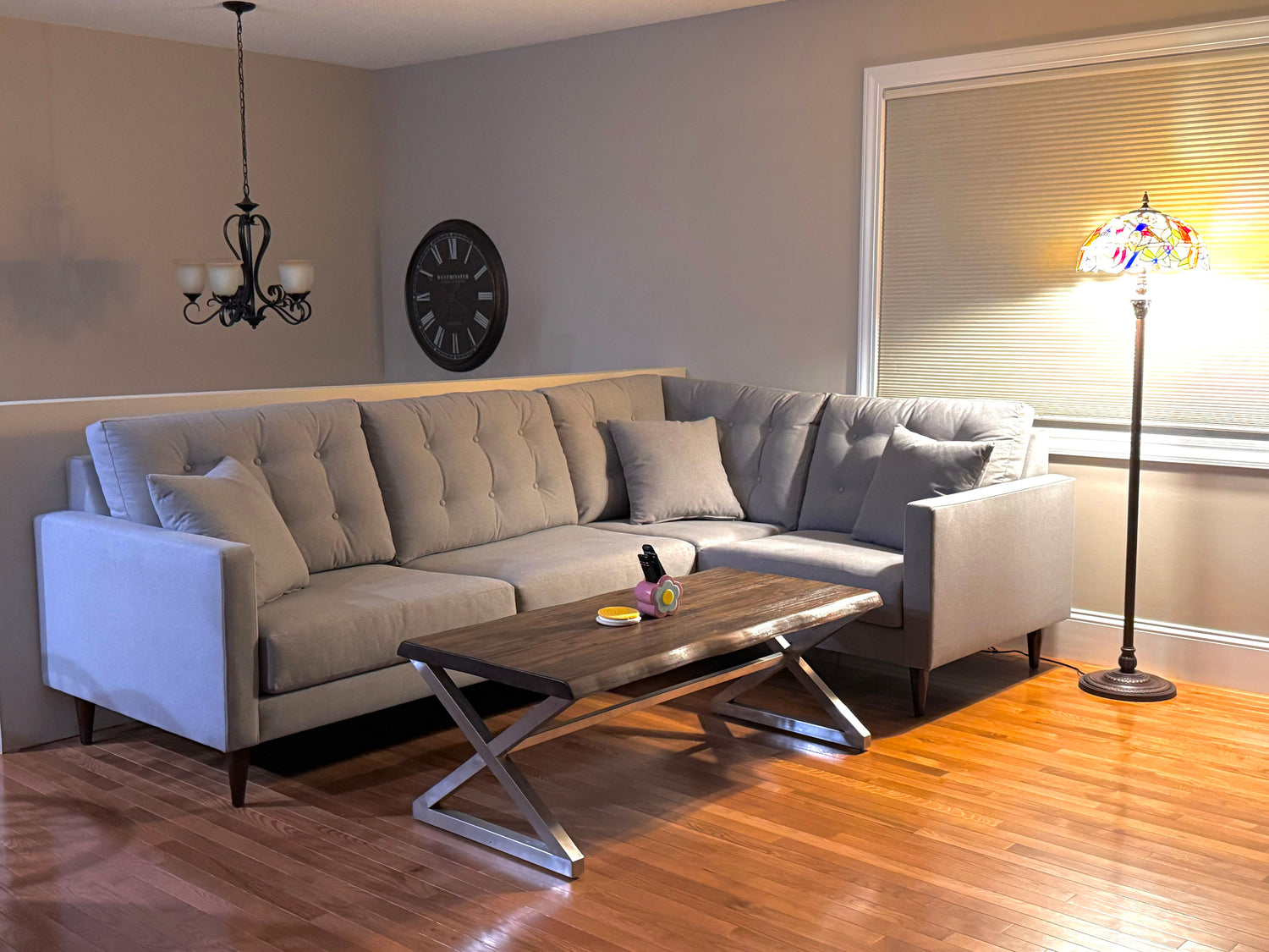 A cozy living room with a gray sectional sofa set against beige walls. A wooden coffee table with metal legs is placed in front of the sofa. The room is lit by a chandelier and a stained-glass floor lamp, and a round clock decorates the wall.