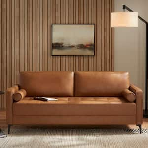 A contemporary living room highlights the StyleWell Goodwin Mid-Century Modern Sofa with Throw Pillows upholstered in brown leather. A backdrop of a wooden slat wall hosts a framed abstract painting, while to the right, a floor lamp with a white shade provides illumination. A side table comfortably holds a book as a neutral rug spans across the floor.