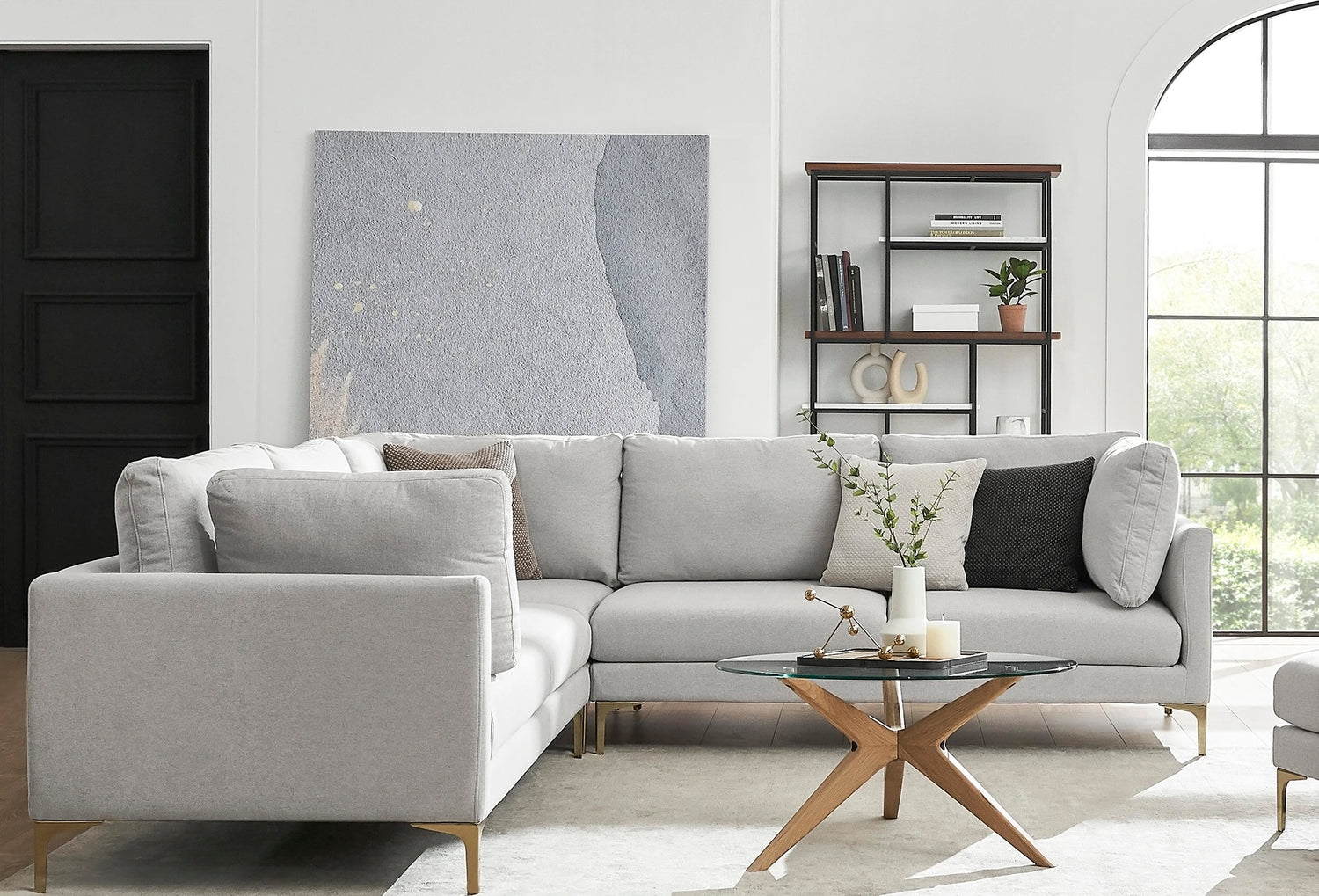 A modern living room featuring a light gray sectional sofa with various cushions, a round glass coffee table with wooden legs, and a tall black shelf holding books and decor. A large abstract painting hangs on the wall, and natural light streams in through nearby windows.