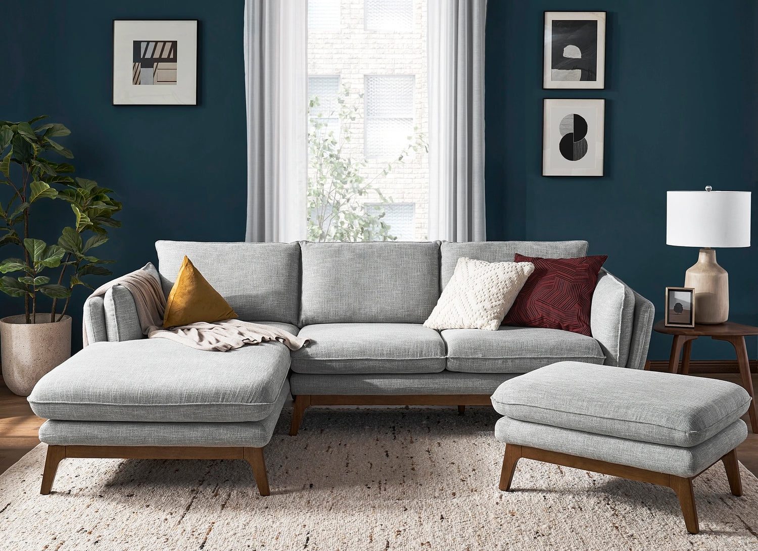 A modern living room with a gray sectional sofa and matching ottoman featuring mid-century wooden legs. The sofa is accented with three throw pillows. Behind the sofa are two framed art pieces and a window with white curtains. A potted plant and table lamp are on either side.