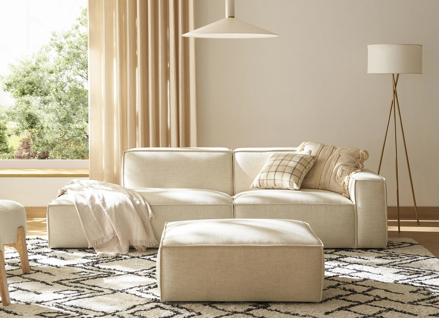 A cozy living room features a beige sofa with pillows and a matching ottoman. A soft blanket is draped over the sofa. The space is adorned with a floor lamp, large window with light curtains, and patterned rug, creating a serene ambiance.