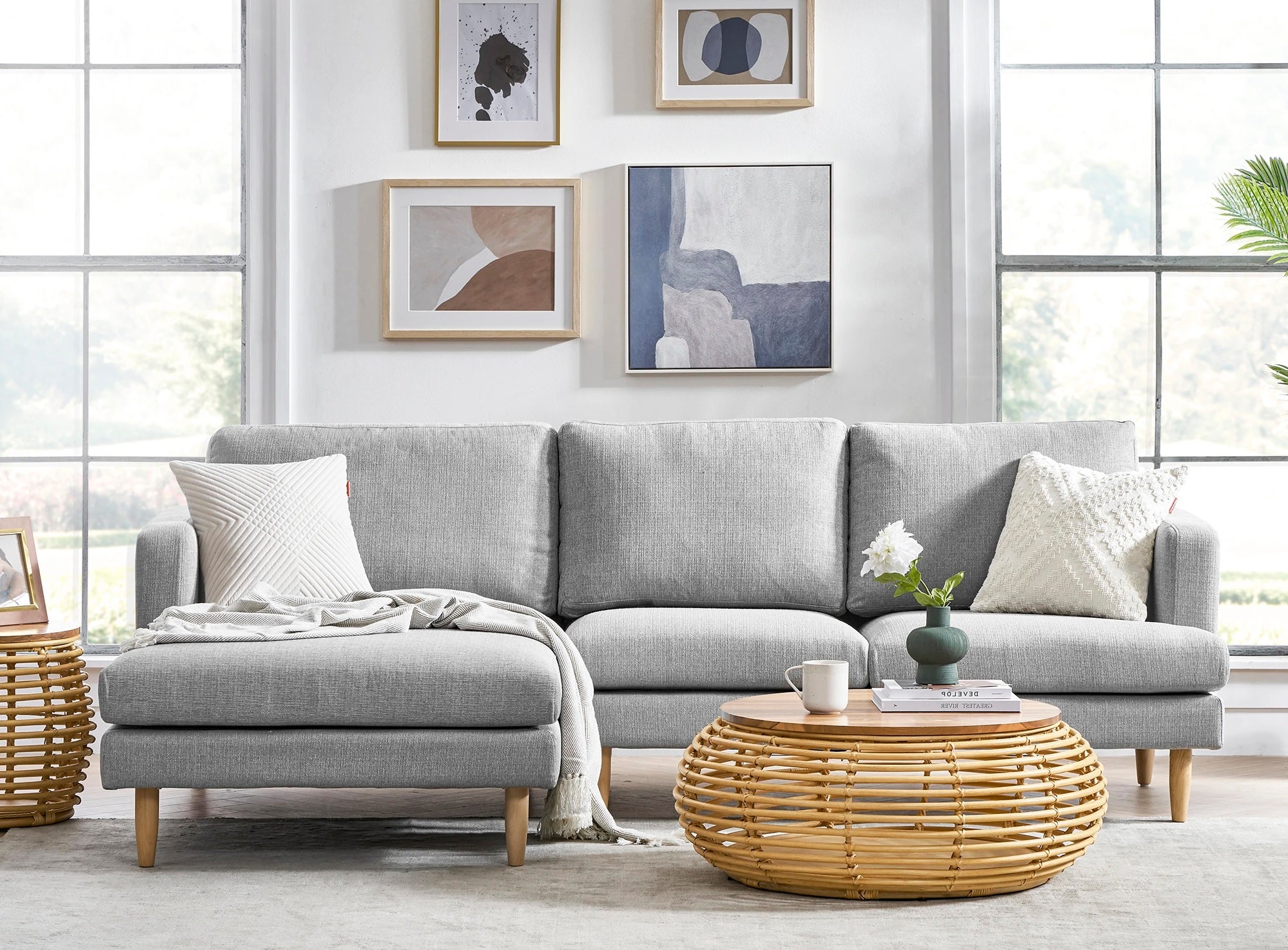 A modern living room with a gray sectional sofa adorned with white pillows and a blanket. In front, there's a round rattan coffee table with a vase of flowers, a candle, and a cup. The walls feature abstract art, and large windows allow ample natural light.