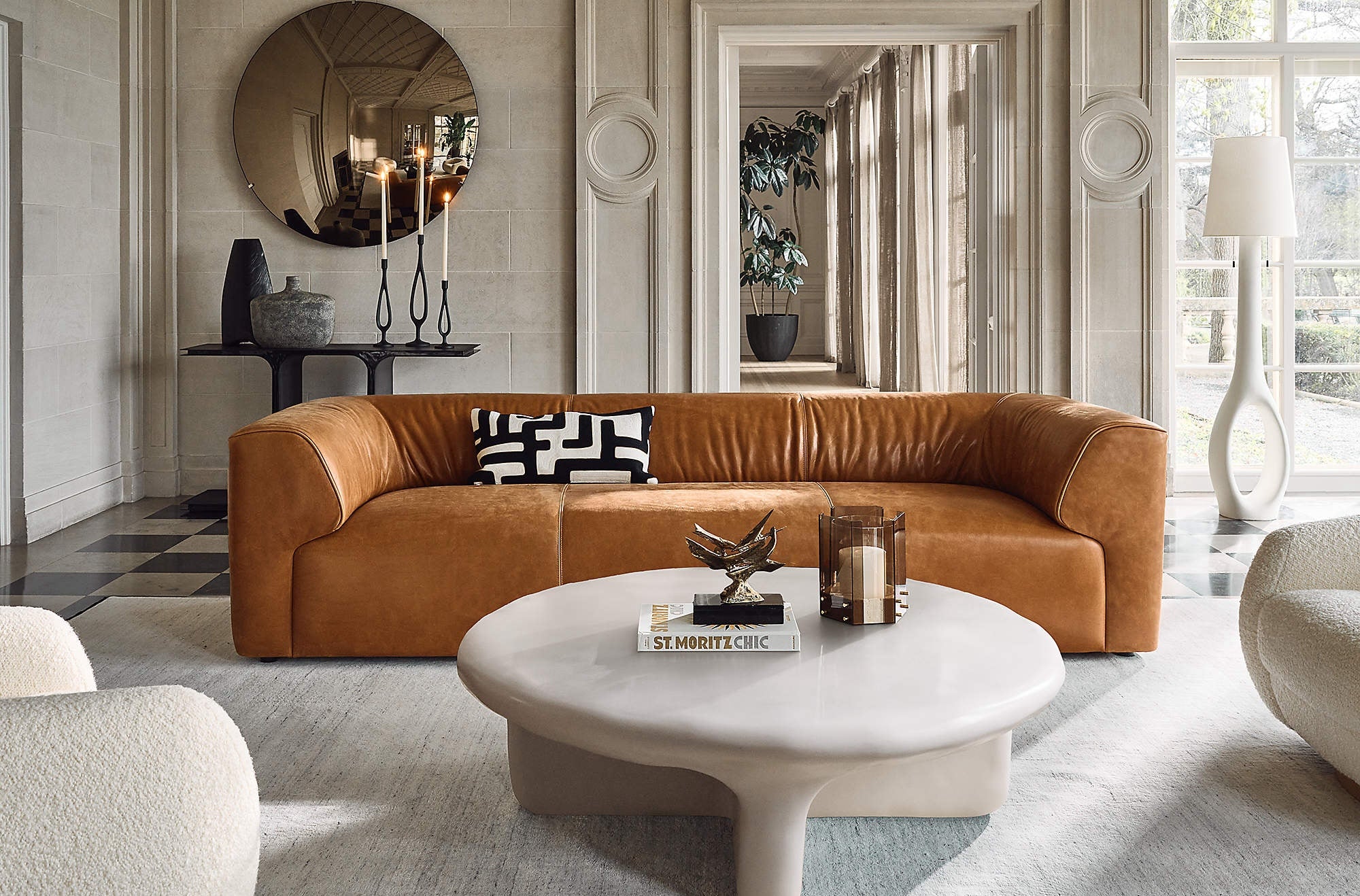 A stylish living room features a caramel-colored leather sofa adorned with a black and white geometric pillow. In front of the sofa is a round, white, modern coffee table. A large, round mirror and decorative items are on the back wall. Natural light streams in from a window.