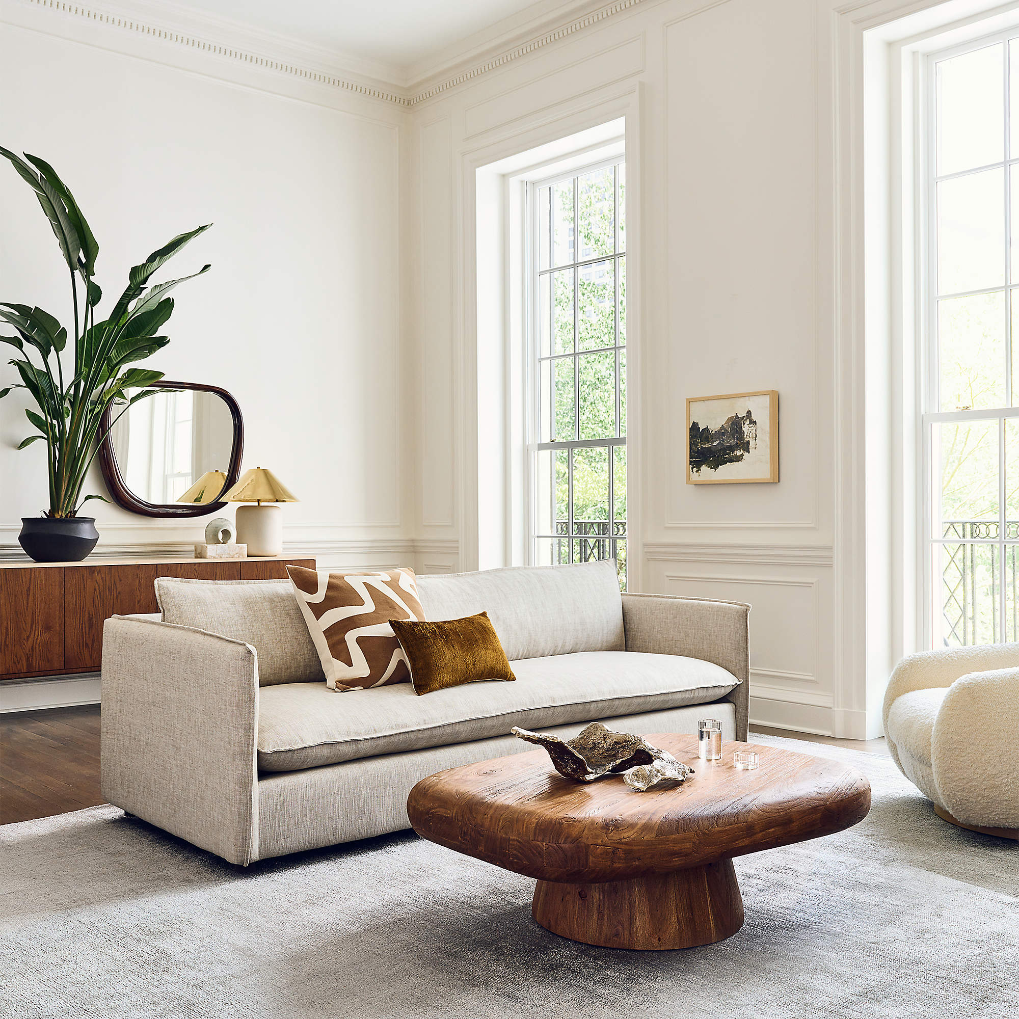 A bright, modern living room features a beige sofa with yellow and white pillows, a wooden coffee table, and a cozy armchair. Large windows allow natural light to flood the space. A wooden console table with plants and a mirror is positioned behind the sofa.