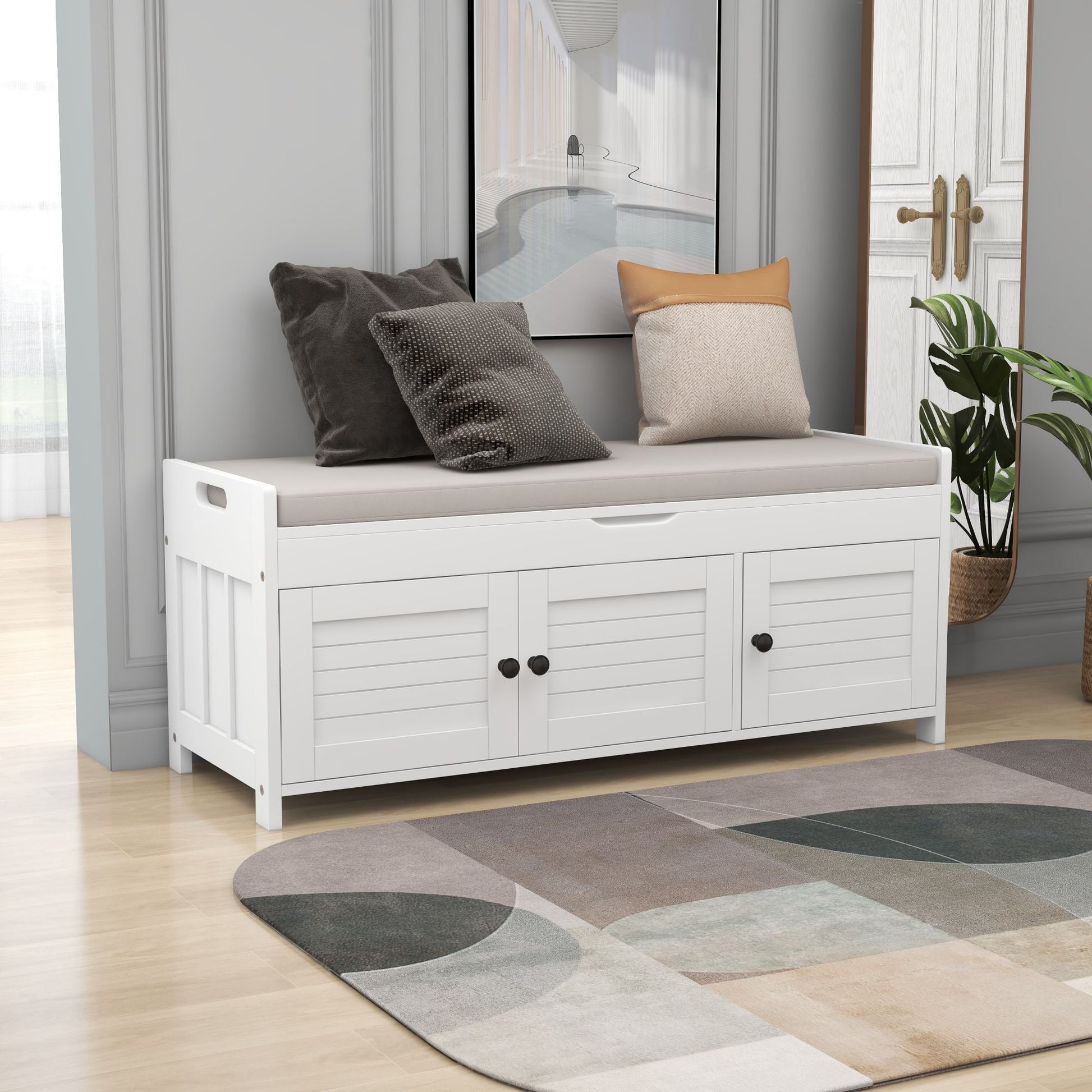 A white wooden storage bench with three shutter-shaped doors, featuring a removable beige cushion and assorted gray and beige pillows. Positioned in a modern room with light wood flooring, an abstract rug, decorative wall molding, nearby wall mirror, and houseplant.