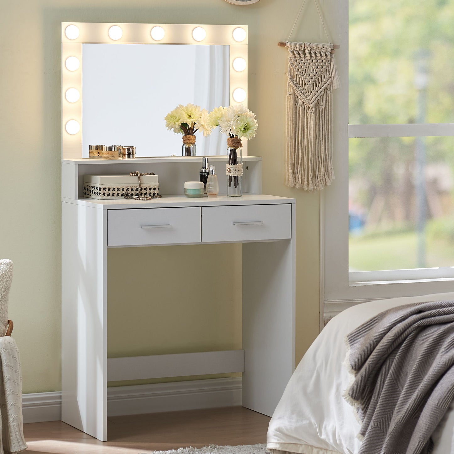 The Vanity Desk With Mirror And Lights features a large drawer and 2-level storage, adorned with makeup, brushes, and two vases of yellow flowers. A macrame wall hanging decorates the background by a sizable window, with part of a bed visible in the foreground.