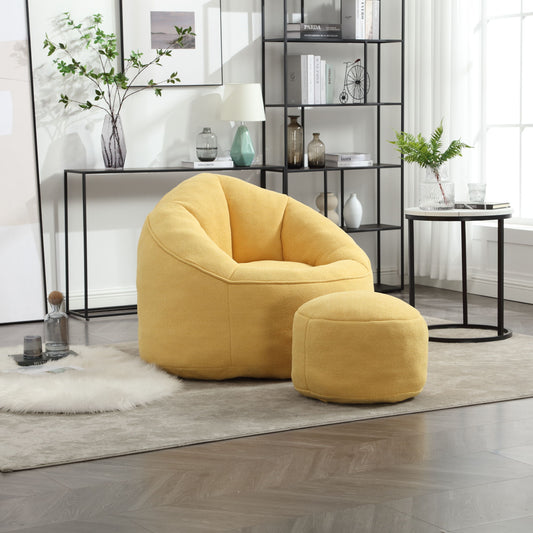 A cozy living room features a Bedding Bean Bag Sofa Chair in yellow with a matching footrest. A black metal shelf with decor, a side table with a plant, and a fluffy white rug complement the space while large windows fill the room with natural light.