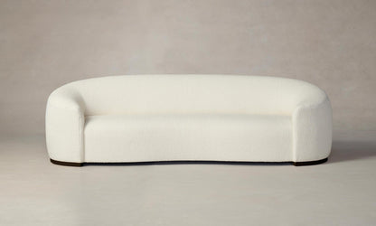 The Chelsea Sofa in Italian Bouclé White, featuring a modern minimalist design with rounded edges and a curved backrest, stands on a neutral-toned floor against a light gray wall. Its plush upholstery offers a soft and inviting appearance.