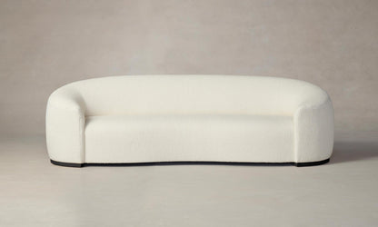 The Chelsea Sofa in Italian Bouclé White showcases a minimalist design with an off-white, curved upholstered form featuring a smooth texture and black base, set against a neutral background for a simple and contemporary look.