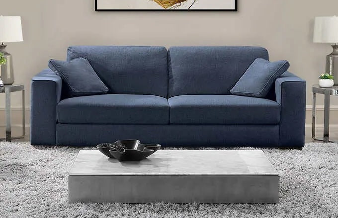 A modern living room features a dark blue sofa with two matching cushions. On either side of the sofa are small tables topped with white lamps. In front of the sofa sits a sleek, rectangular coffee table on a light gray shag rug.