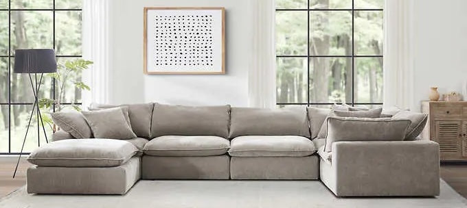 A spacious, neutral-toned sectional sofa is centered in a modern, well-lit living room with large windows. A black floor lamp stands to the left, and a minimalist framed artwork hangs above the sofa. The wall on the right has a wooden cabinet with decor items.