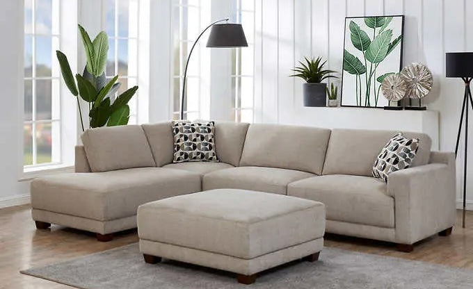 A modern living room with a beige sectional sofa and matching ottoman. The sofa has decorative black and white patterned cushions. Behind the sofa, there are two large potted plants, a floor lamp, and a framed botanical artwork. Large windows provide natural light.