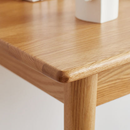 Close-up of the Computer Desk in Oak Natural, highlighting its smooth, rounded edge and light wood grain. Part of the table leg is visible along with a blurred white object on the tabletop.