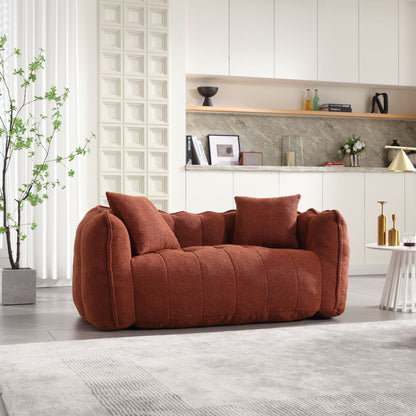 A cozy, textured rust-colored beanbag chair with a high resilience foam core for two people sits in a modern living room. Behind it, a marble backsplash and minimalist shelves with decor adorn white cabinets. A potted plant adds greenery beside floor-length curtains.