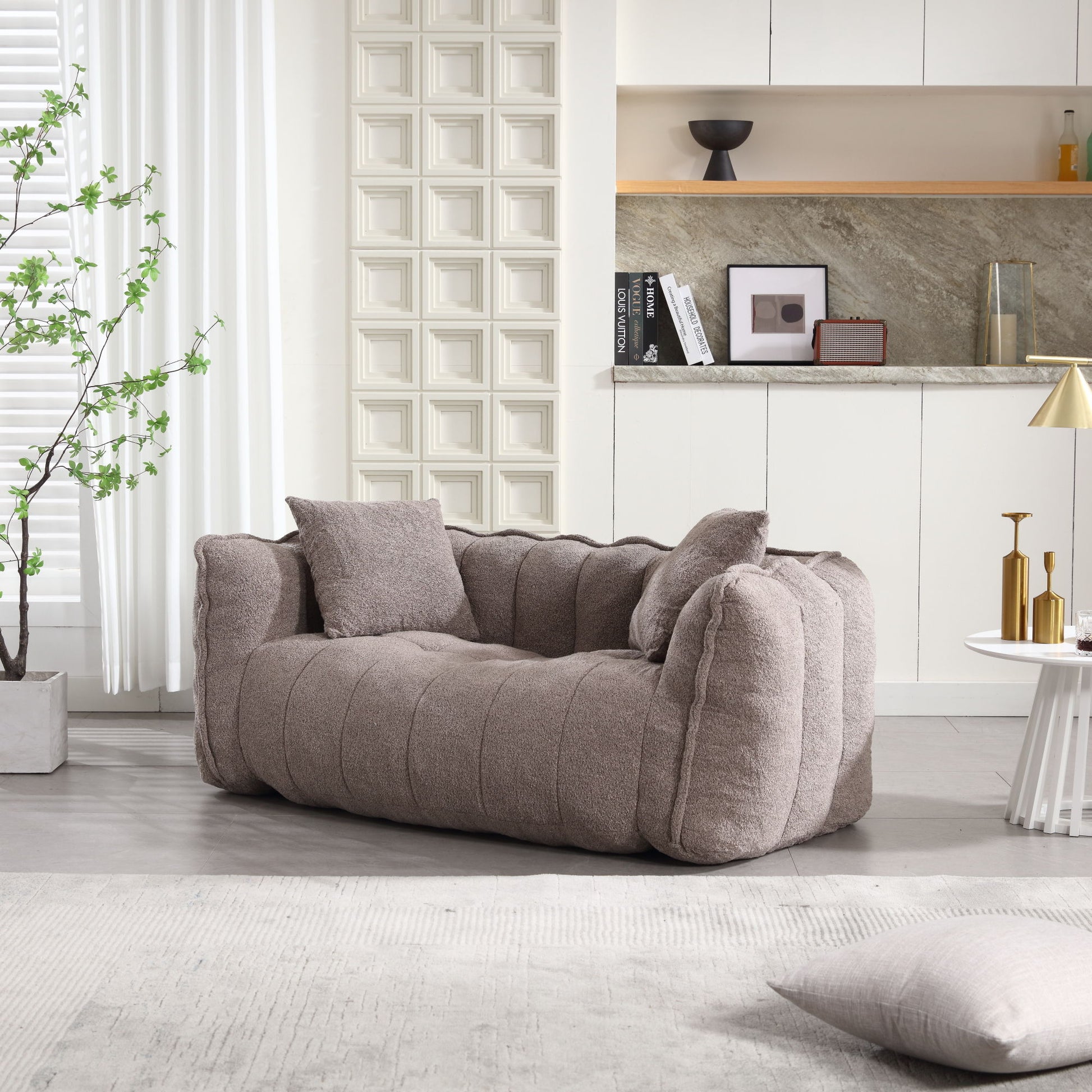 A modern living room features a plush taupe sofa, and in the foreground is a Soft Beanbag Chair with high resilience foam core for two people. Behind, there's a decorative wall with square patterns, white cabinets, and a stone countertop. A small potted plant graces the round coffee table filled with books and a vase as natural light bathes the space.